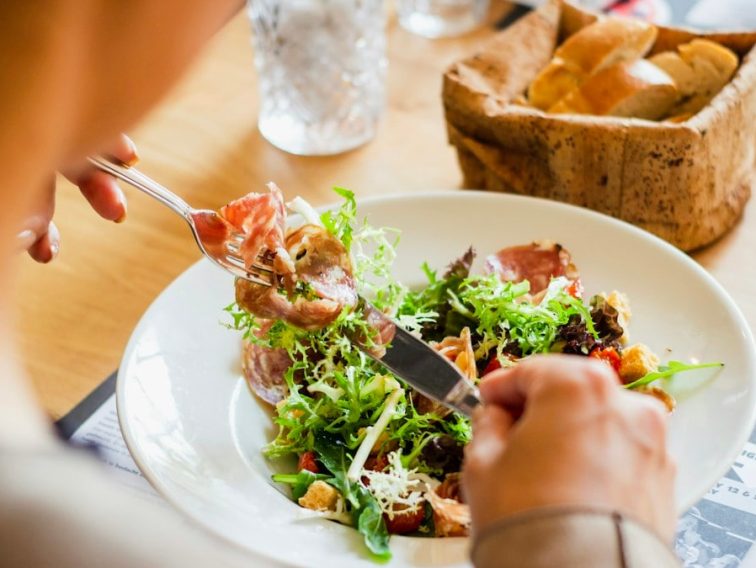 A person eating a salad with cured meat and croutons from a white plate