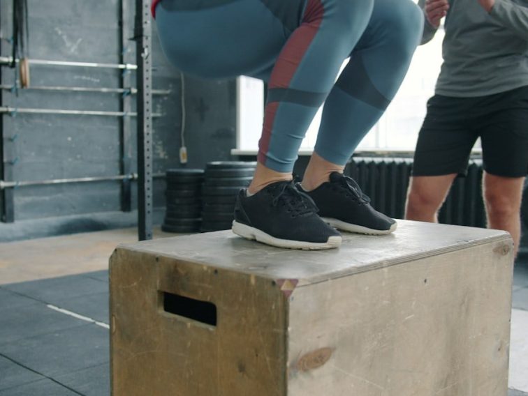Low shot of sportswoman jumping onto wooden box during sports training with coach while man standing and watching. People, lifestyle and fitness concept.